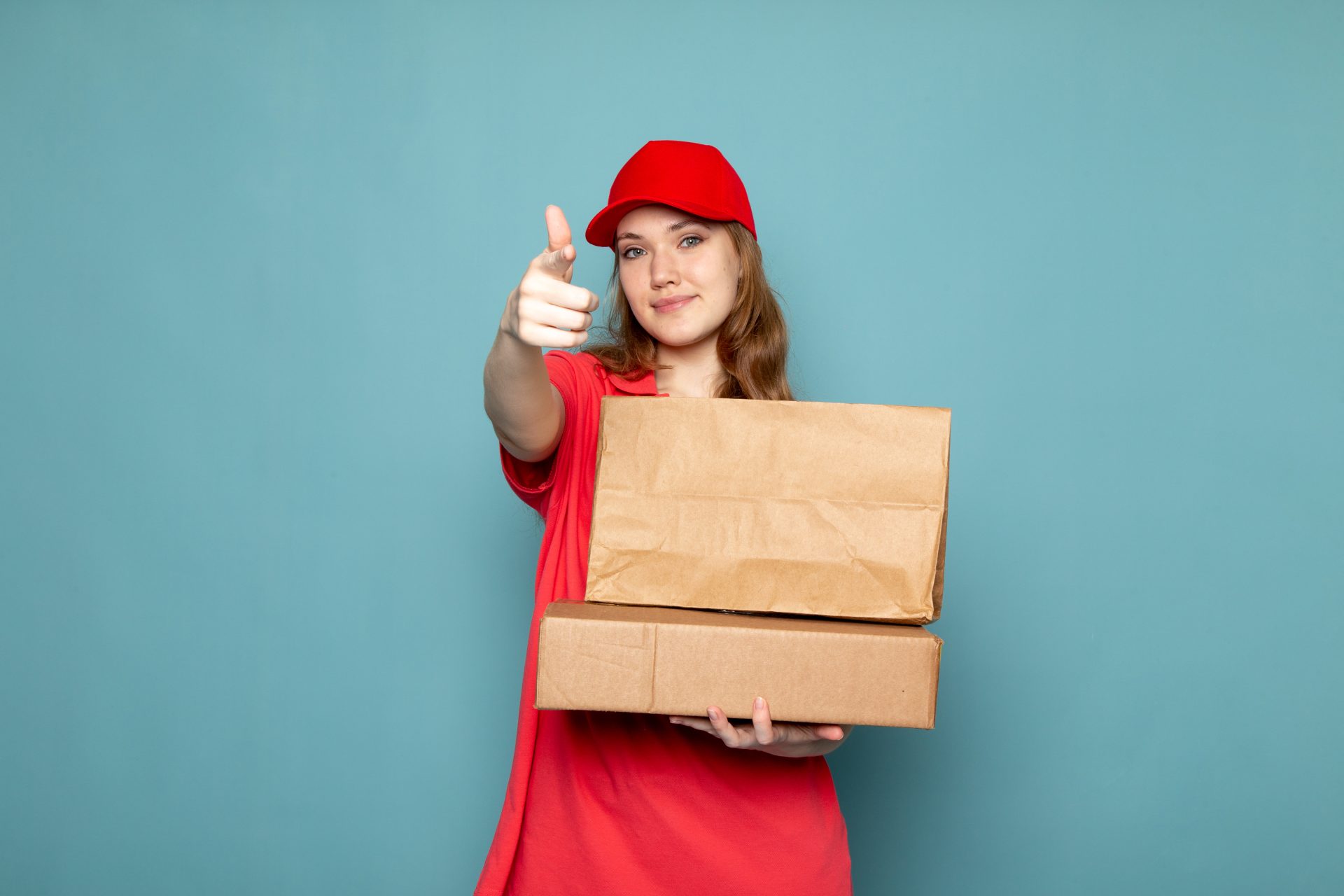front view female attractive courier red polo shirt red cap holding brown packages smiling posing blue background food service job